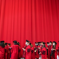The image captures a group of graduates, standing against a vibrant red curtain backdrop
