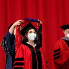 A young woman wearing a black graduation cap, glasses, and a white face mask is standing on a stage in front of a red curtain