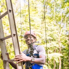 A young man, wearing a red helmet and a safety harness, is ascending a metal ladder in a ropes course set high amongst the trees