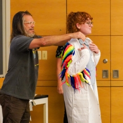 a man with long gray hair and a beard is adjusting the collar of a white lab coat worn by a young woman with red curly hair and glasses