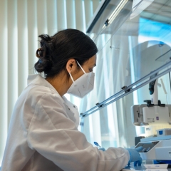 a female scientist working in a laboratory. She is wearing a white lab coat, blue nitrile gloves, and a white N95 face mask